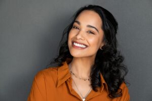 A young woman with long dark curly hair smiles warmly at the camera. She is wearing an orange top and a delicate necklace, standing in front of a plain gray background.